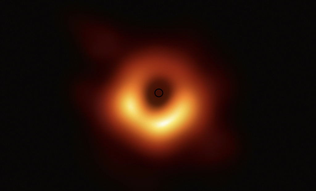 Sur un fond noir, il y a une auréole jaune-orangée brillante autour d’un centre noir. Au centre de ce centre noir se trouve un petit cercle noir. L’auréole est plus épaisse et brillante dans la partie basse de l’image.