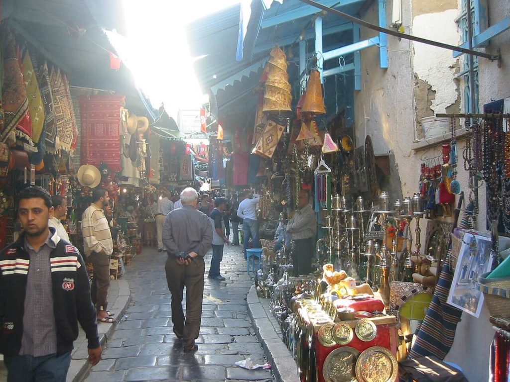 Photographie d’un souk de la médina de Tunis : l’endroit est très fréquenté, des tissus, objets d’arts, tasses et tapis sont entassés de chaque côté de la rue pavée. Les commerçants se trouvent devant leurs produits.