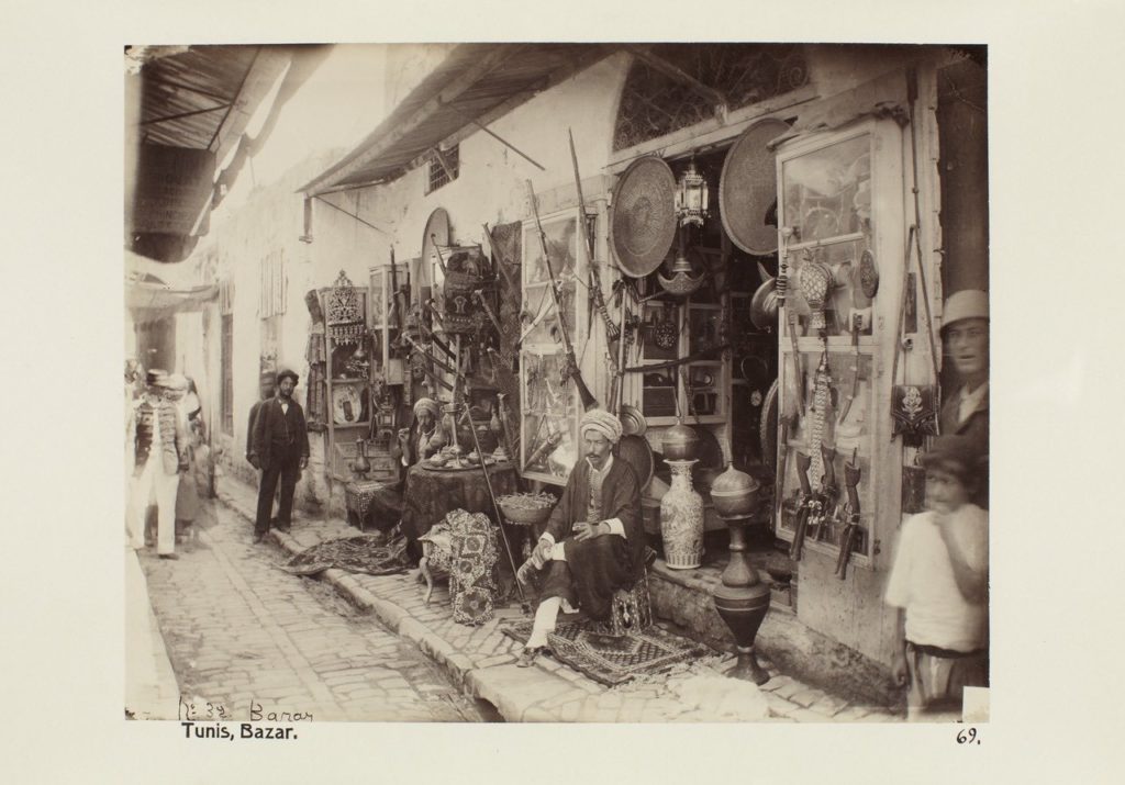 Photographie sépia d’un souk. Dans une rue, un commerçant vêtu d’un turban est assis devant de nombreux objets (tissus, vases, tasses et lanternes). Des personnes passent dans la rue pavée en arrière-plan.