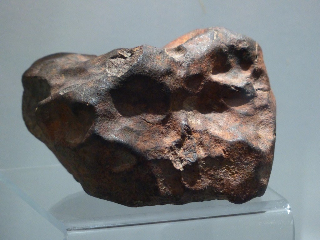 Photographie d'une météorite de couleur noir-brun, posée sur une table transparente. La météorite est parsemée de nombreuses crevasses arrondies.
